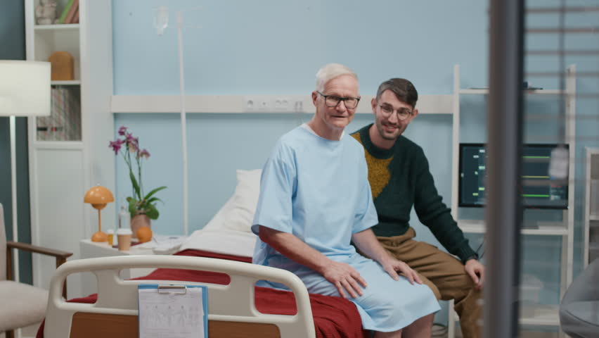 Slow motion of little girl supporting grandpa with drawing and embracing him while visiting in hospital room with father