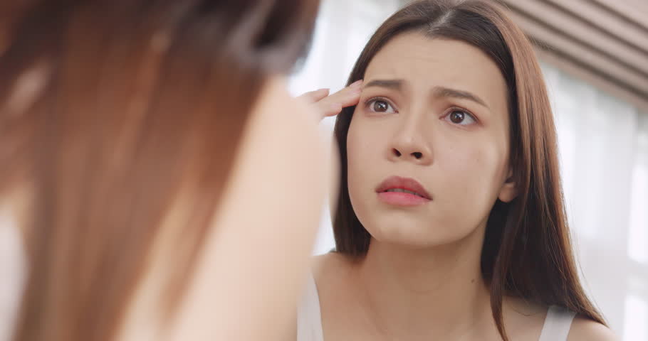 Young woman worried about the wrinkles on her face while looking in the mirror at home.