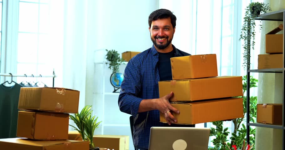 Indian Asian small businessman standing proudly in a store room office, happily showing a pile of cardboard carton boxes arranged for shipment, dispatch, and delivery after successful order packaging