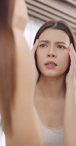 Vertical clip. Young woman worried about the wrinkles on her face while looking in the mirror at home.