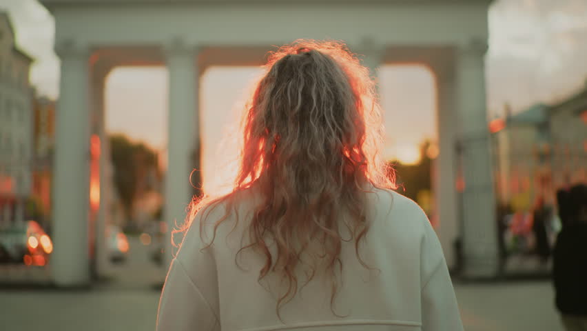 back view of woman with curly hair walking toward sunlit archway in urban setting as glowing red sunlight illuminates her hair with blurred motion of cars and people in background