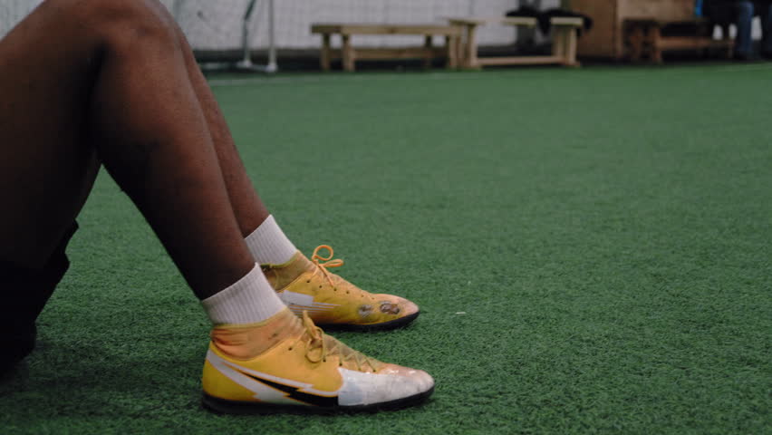 A pan left shot from a footballer’s yellow boots to an African footballer wearing a purple bandana lying on the grass after a tough training session.