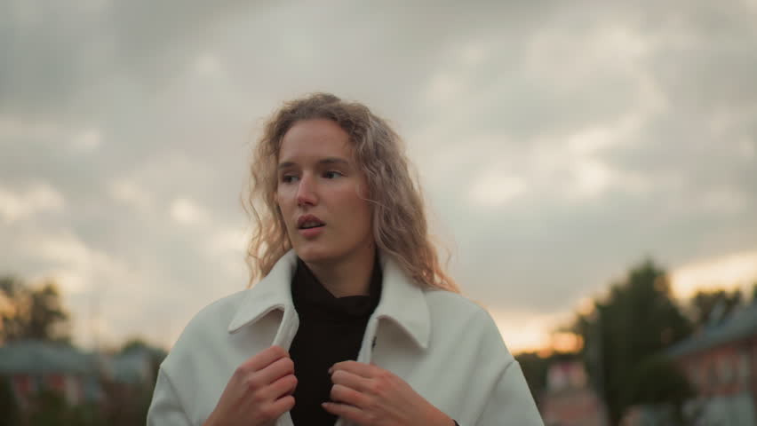Young girl feeling cold pulling white coat tightly around herself under cloudy sky during chilly weather, with soft blurred background of green trees, buildings, and evening natural light