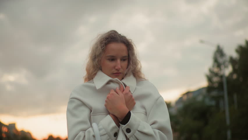 Young girl feeling cold pulling white coat tightly around herself under cloudy sky during chilly weather, with soft blurred background of green trees, buildings, and evening natural light