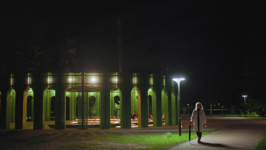 Young girl dressed in white coat walking through serene night park holding umbrella, passing by glowing streetlights and modern illuminated green pavilion structure with people in background