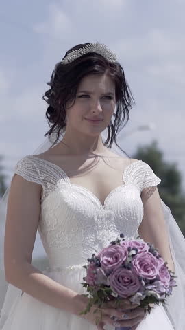 slow motion young pretty woman in wedding dress with cleavage holds purple bouquet against town buildings