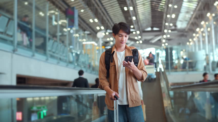 Happy asian man with backpack checking flight in phone on escalator in airport terminal, People concentrating typing digital device for travel in transportation, Concept of travel
