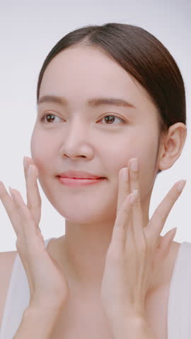 Vertical clip. Beautiful young woman applies moisturizer to her face on white background.
