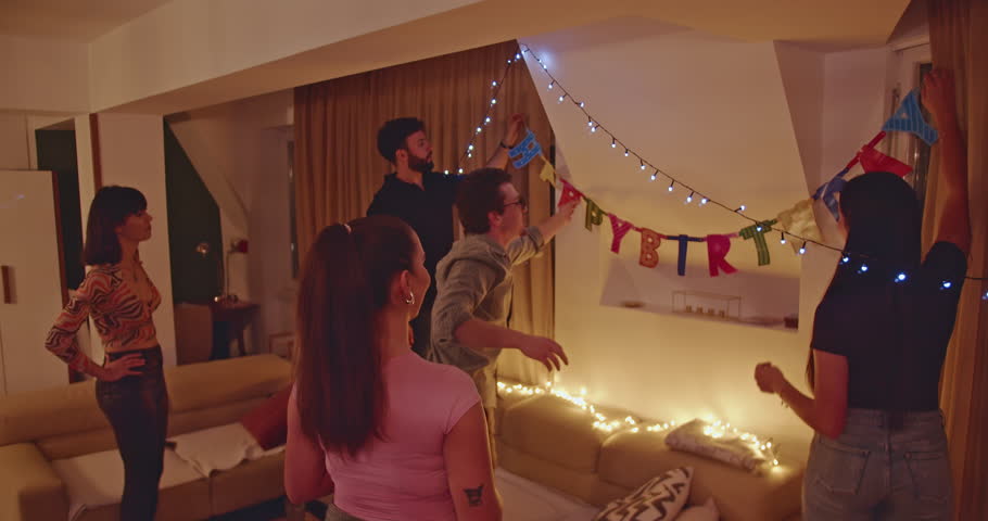 Friends decorating their home for a birthday party. They are hanging up a Happy Birthday banner and string lights. The friends are excited and happy to be celebrating together.