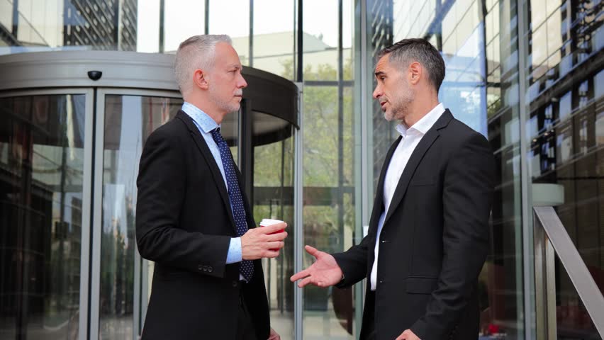 Two business professionals engaged in an intense discussion while analyzing information on their smartphones with coffee in hand outside a corporate building during a busy workday