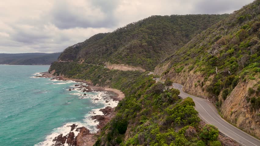 Aerial view of a car driving along the winding Great Ocean Road, surrounded by lush hills and turquoise ocean