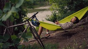 Man travels on bicycle, relaxing in green hammock, surfing Internet on smartphone, listening music on headphones in forest near lake. Cyclist in hammock at campsite by river. Male on bike in hammock. - Powered by Shutterstock - Get 15% off with code: PIKWIZARD15