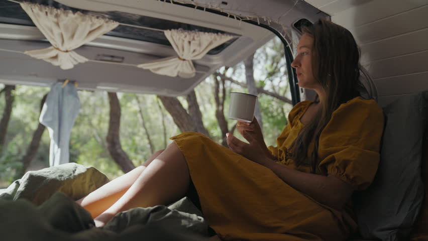 Youn adult woman enjoying a peaceful morning with a cup of coffee while relaxing inside a van amidst lush trees