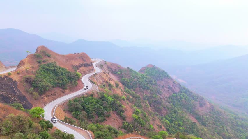 Aerial view bendy trail cuts craggy hill under hazy sky, Drone shot zigzag road above deep valley, Vehicle moves on mountain track, Over ridged terrain and trees, Panorama show slant route by forest