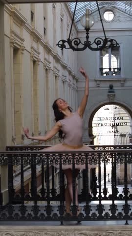 Ballet dancer in a soft pink tutu poses gracefully on pointe inside a vintage shopping gallery with ornate architecture and arches. 