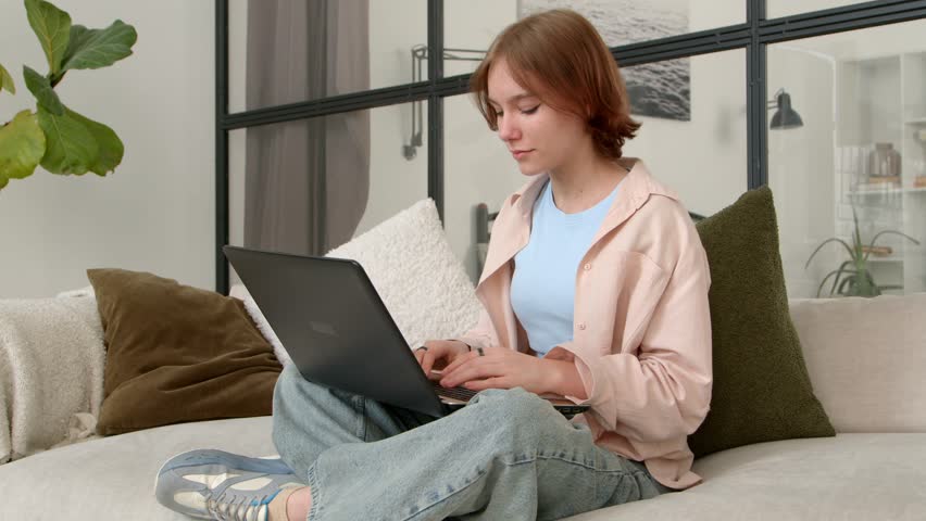 Adorable teenage girl sitting cross-legged on sofa, networking and online messaging in social media using laptop, being surprised and shocked by latest rumors while spending leisure at home.