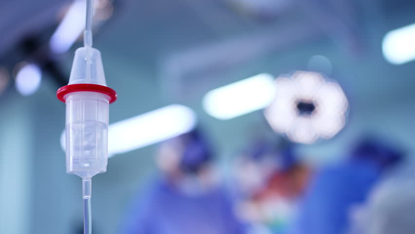 Surgical procedure with IV drip in focus. An IV drip is visible in a surgical room as medical staff conduct a procedure in a sterile environment.