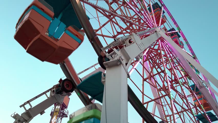 Colorful Ferris wheel seen from below as it rotates under the blue sky at a fairground in Cuernavaca, Morelos, Mexico. Metal structure and gondolas reflect sunlight in motion
