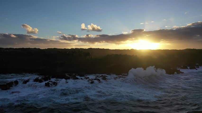 Powerful waves crash into the dark volcanic coastline of Hawaii as the sun sets behind the hills, casting warm light across the ocean and sky.