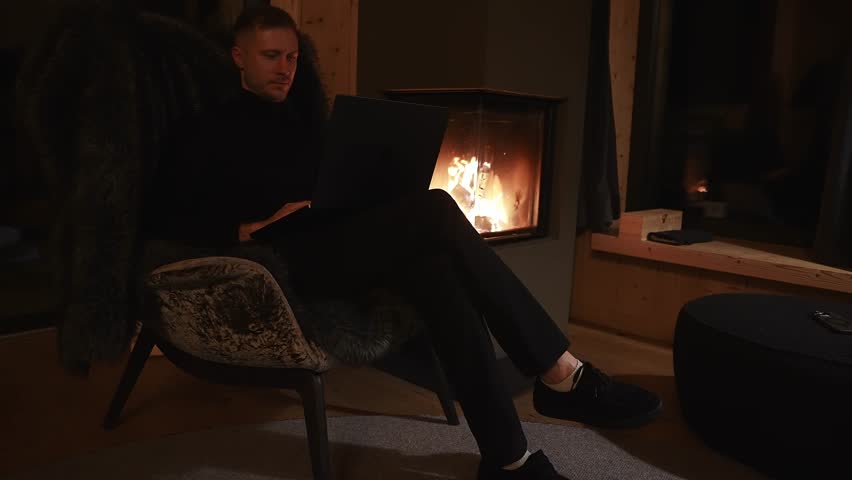 A man sits in a plush chair with a fur throw, working on a laptop near a glowing fireplace. Wooden walls and large windows suggest an alpine setting.