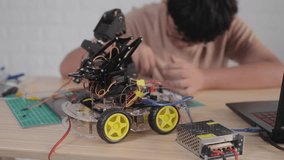 Close-up view of a robotic car with an attached mechanical arm, wired and connected to components, while an Asian teenage boy works in the background on a STEM engineering project at home. - Powered by Shutterstock - Get 15% off with code: PIKWIZARD15