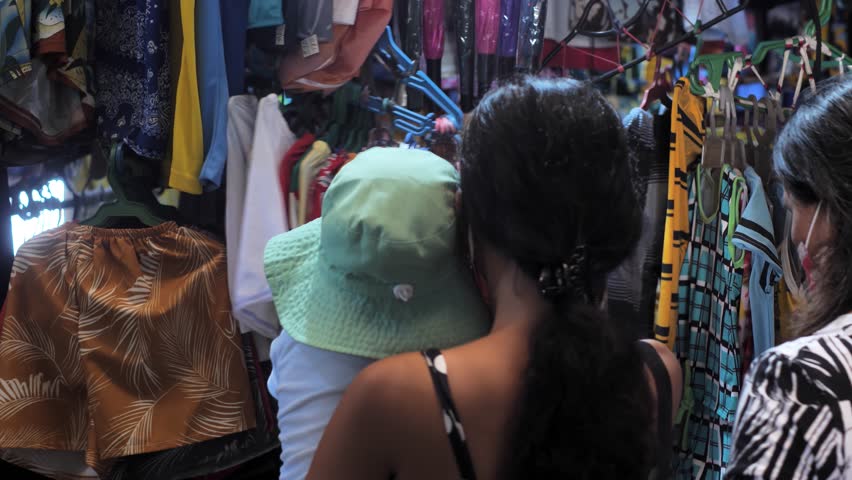 Brunette woman with baby in arms chooses outfit on outdoor market display rear view. Young mother with child in panama visits street shop in Dumaguete