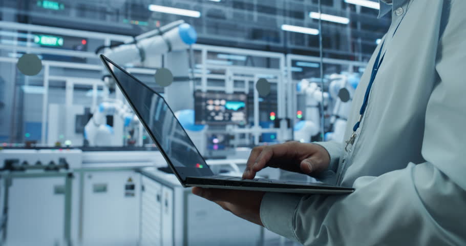Electronics Factory Manager Working on Laptop at an Industrial Manufacture with Robotic Arms. Anonymous Male Specialist Monitoring Data on a Computer, Checking and Planning Production Process