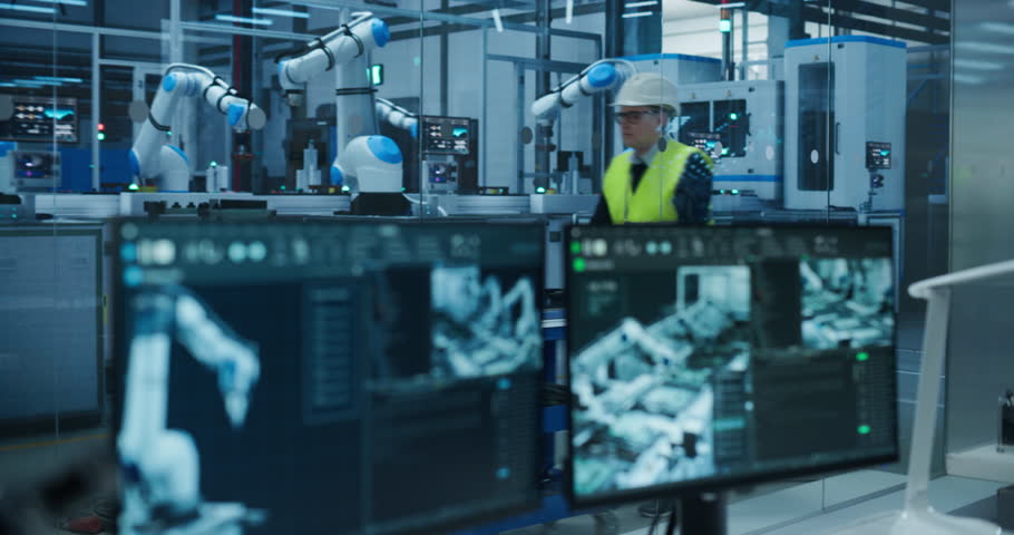 Female Engineer Sitting Behind a Desk in a Modern Product Development Facility, Using Industrial Computer Software on Dual Screens, Monitoring Electronics Production Line with Robotic Arms