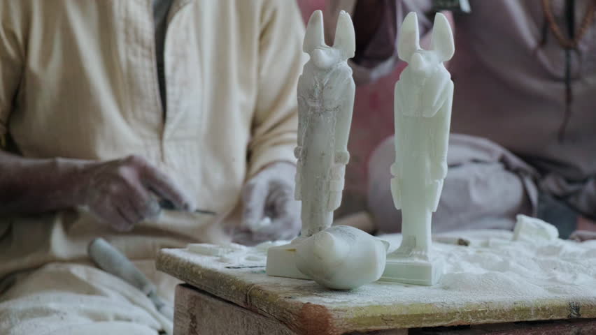 Unrecognizable craftsman is making traditional ancient egyptian trinkets. Figurines of Anubis at foreground. Alabaster carving workshop in Luxor, Egypt