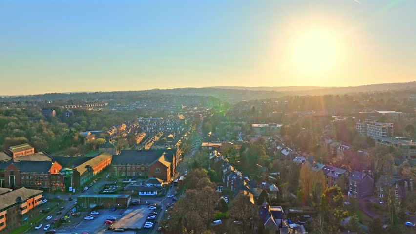 A high drone shot reveals Sheffield's urban sprawl, sun rising over distant hills in morning light. Autumn tones, blue skies, and flowing traffic create a vibrant and serene cityscape.