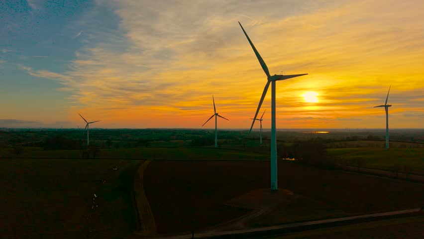 Electric windmill spinning and producing eco energy at sunset. Wind turbine moving energy capturing blades to produce clean output under dramatic orange evening sky. Moving circular blades supporting