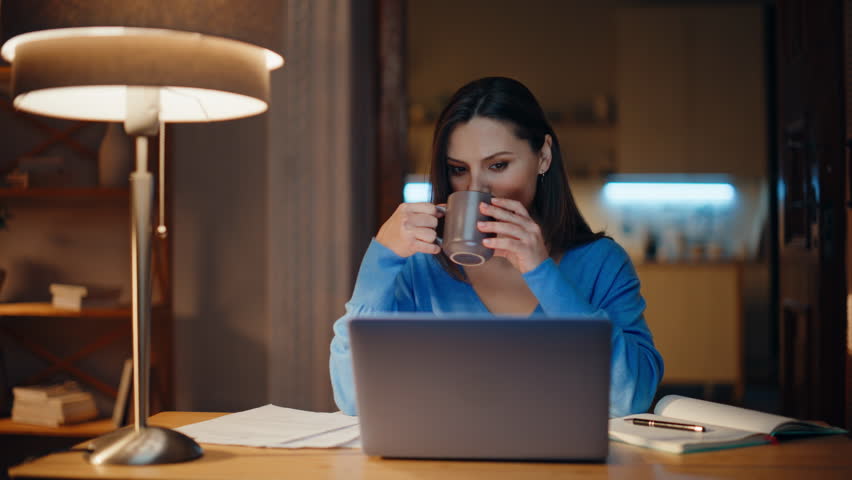 Relaxed freelancer drinking coffee at remote computer workspace. Startuper creating project at evening apartment looking laptop closeup. Businesswoman writing ideas in copybook sitting dark workplace.