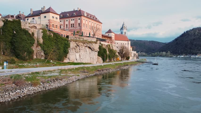 Aerial drone view of Dürnstein in the Wachau Valley in Lower Austria, showcasing terraced vineyards, Danube River, historic medieval villages, lush landscapes of UNESCO World Heritage during sunset.
