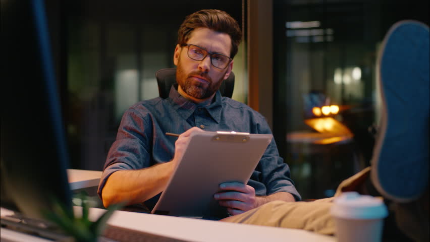 Thoughtful male businessman Caucasian business man guy in dark evening office relaxed thinking looking at computer write notes deal document paperwork task writing brainstorm ideas planning night job