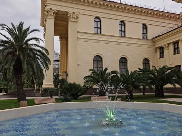 Fountain with a green flower in the middle. The fountain is surrounded by trees and benches. The building behind the fountain is white in Sochi