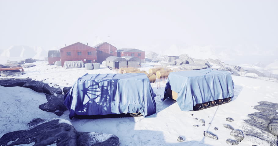 A remote winter landscape features red buildings surrounded by snow and ice. Two carts covered in blue tarps sit nearby, with rocky terrain and distant mountains in the background.