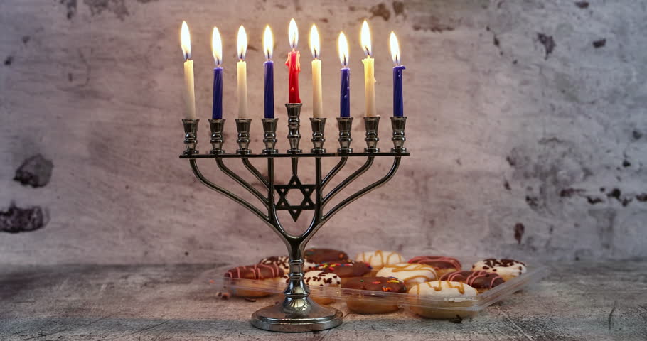 Silver menorah with nine burning candles, red center candle, placed on textured surface with decorated pastries in foreground. Star of David visible in menorah design, textured concrete wall