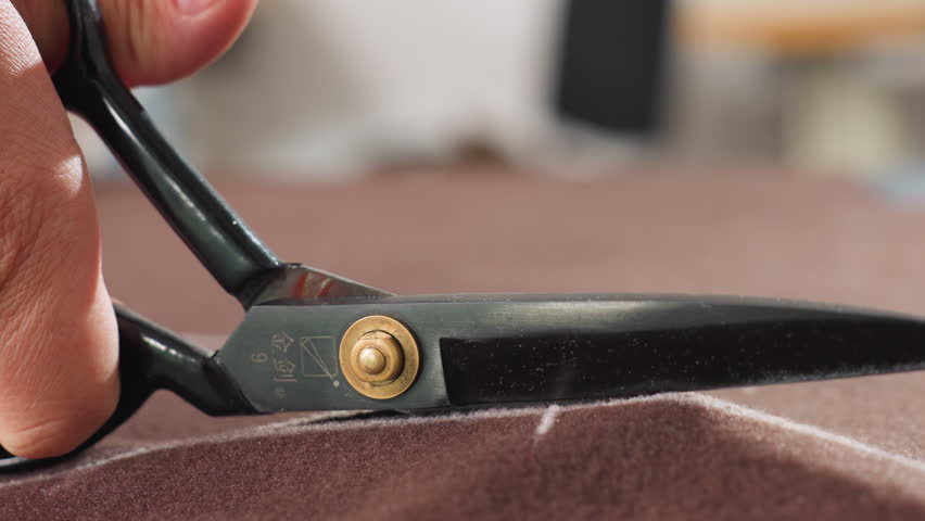 Close up view of hand gripping black scissors cutting brown textile along chalk markings on work surface, precision tailoring process captured in workshop setting, focus on blade slicing fabric