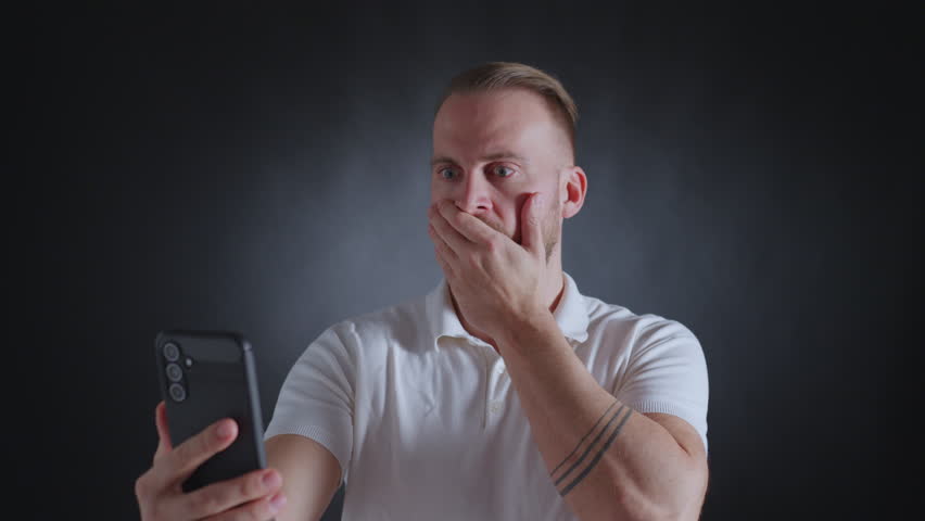 Man in white shirt, shocked expression, covering mouth, holding phone, tattoo on arm, dark background.
