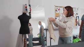 Seamstress lifts beige tailored vest toward black dress form, aligning shoulders before careful pinning, evaluating drape and structure in studio filled with patterns, color swatches, board behind - Powered by Shutterstock - Get 15% off with code: PIKWIZARD15