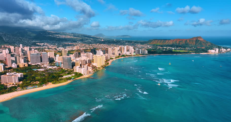 Slow waves with foamy rims roll to the shore. Drone footage high above the waterscape of the Pacific Ocean. Waikiki resort hotels at the coastline. Diamond Head Carter at backdrop.