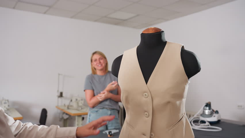 Clothing designer places hands on beige sleeveless jacket worn by mannequin while smiling and laughing joyfully with partner in shared moment of celebration after completing fashion project together