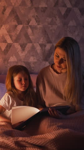 Careful long haired mother reads fairy tale to cute little daughter sitting together on bed with pink plaid in semidark children room slow motion