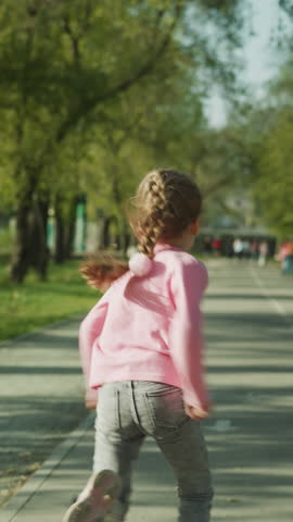 Playful little girl with braided fair hair runs along empty asphalt pedestrian road in spring park backside view. Active sportive child has fun slow motion