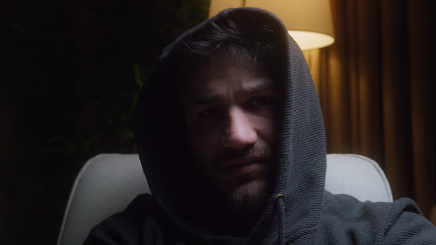 Confused young man wearing a hoodie, sitting on a couch in a dimly lit room, frowning and giving a strange look while a warm floor lamp glows softly in the background