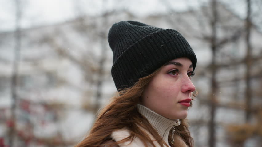 Close up side shot of young girl in black cap and brown jacket walking with serious expression and wind-blown hair against blurred city background under cold overcast sky during quiet winter day
