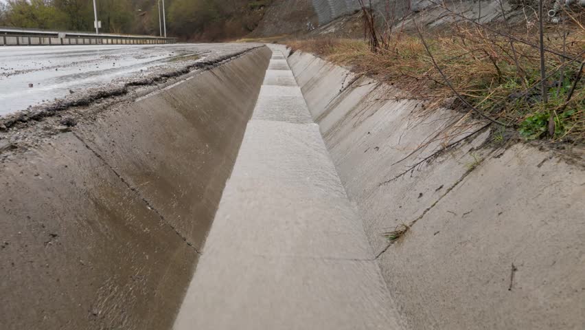 Rainwater streaming down concrete drainage channel alongside wet asphalt roadway