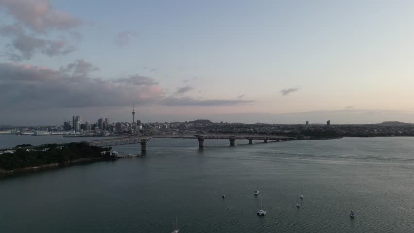 Drone shot at dusk showing Auckland’s skyline in the distance with the Harbour Bridge, boats on the water, and soft sunset light over the Waitemata Harbour.
