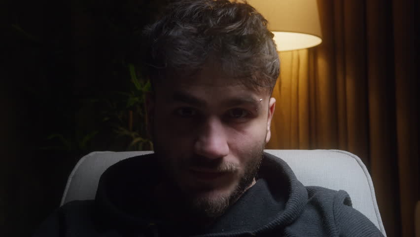 Young Man Sitting in Dimly Lit Room Pointing at Camera with Serious Expression