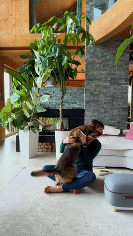 VERTICAL: Woman laughing heartily as she plays with her shepherd dog on a cozy living room rug. Moment of authentic joy and connection in a thriving home indoor jungle is bathed in soft daylight.